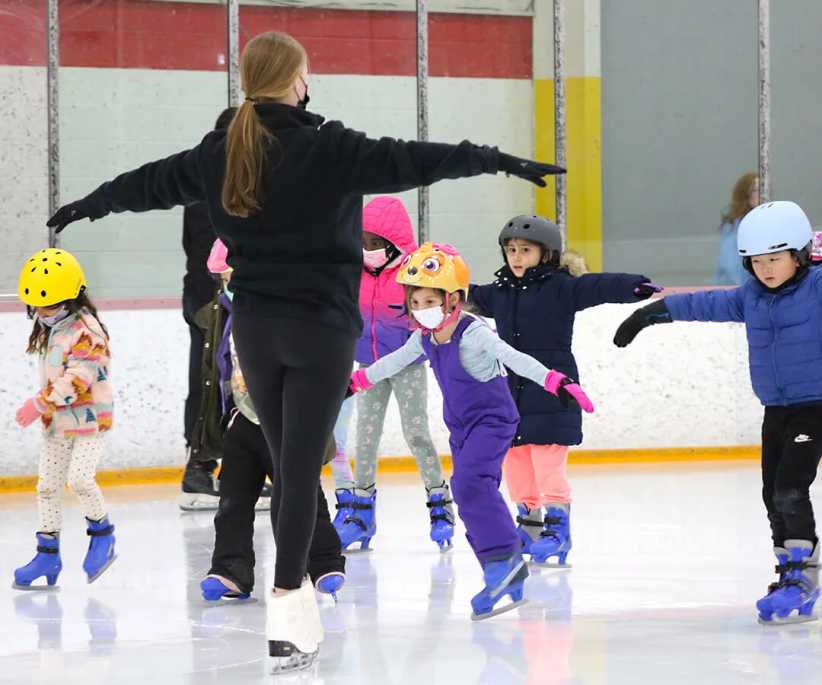 An instructor guides the children on an ice rink as they practice skating with arms outstretched, wearing helmets, gloves, and winter clothes.