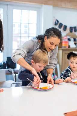 A young boy eats orange food with a spoon, assisted by a woman. He is seated at a table in a bright kitchen with other children.