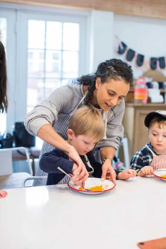 A young boy eats orange food with a spoon, assisted by a woman. He is seated at a table in a bright kitchen with other children.