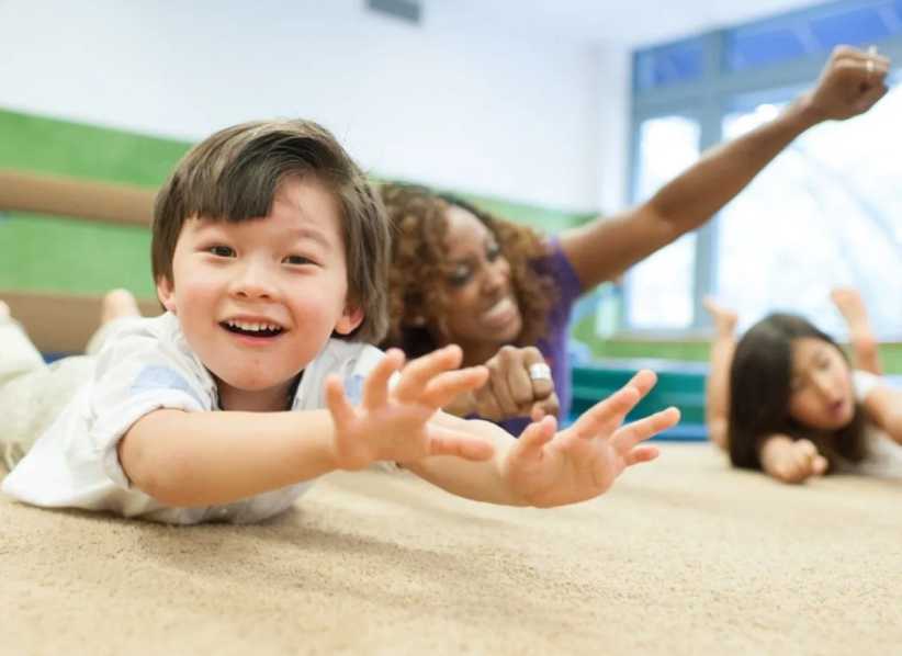 Children lie on the floor reaching forward with their arms, participating in a playful classroom activity in a bright, indoor setting.