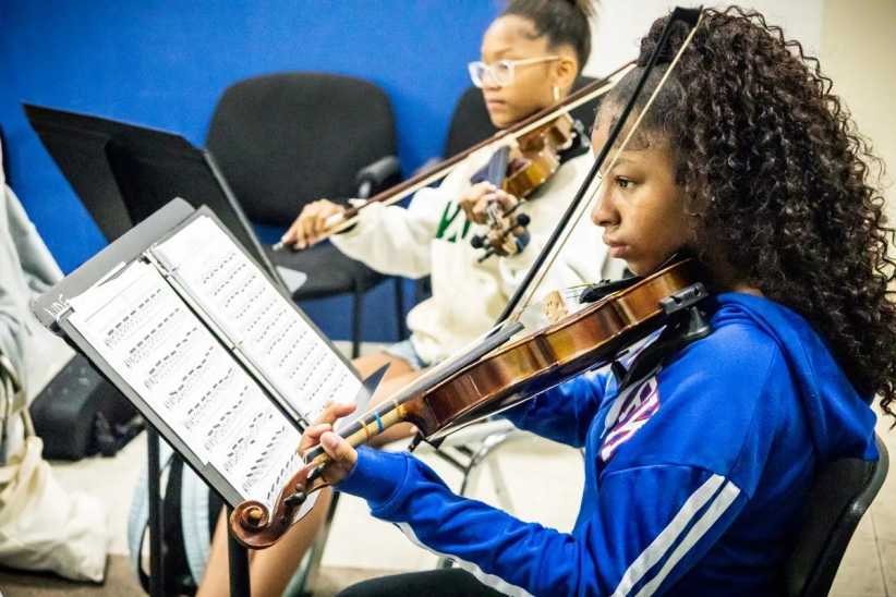 Two students play violins in a classroom, reading sheet music on stands, with chairs and a blue wall seen in the background.