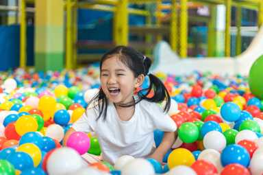 A young girl with pigtails laughs and plays in a colorful ball pit at an indoor playground, surrounded by red, blue, yellow, and white balls.