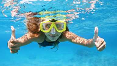 A swimmer wearing yellow goggles gives two thumbs up underwater in bright blue ocean water, with sunlight creating patterns on her skin.