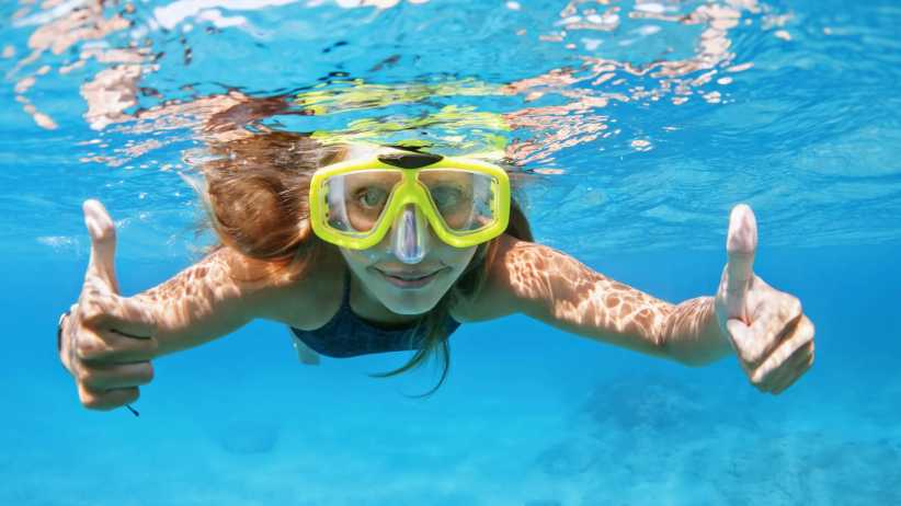 A swimmer wearing yellow goggles gives two thumbs up underwater in bright blue ocean water, with sunlight creating patterns on her skin.