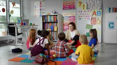 Children sit in a circle around a teacher on a colorful mat in a bright classroom, listening and engaging in a group activity.