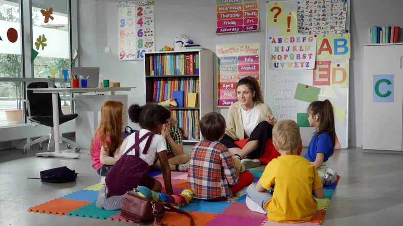 Children sit in a circle around a teacher on a colorful mat in a bright classroom, listening and engaging in a group activity.