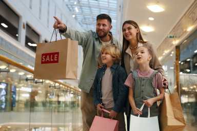 A smiling family of four stands in a shopping mall, holding shopping bags with a "Sale" sign, as the father points excitedly ahead.