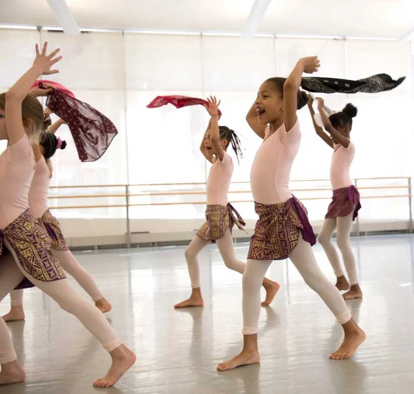 Girls dancing joyfully in a bright studio, wearing pink leotards and patterned skirts, waving colorful scarves above their heads.