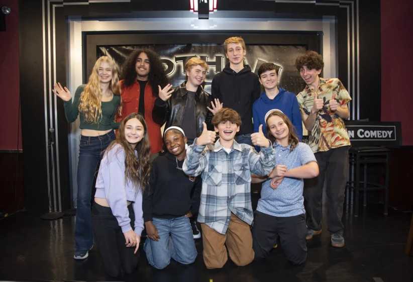 Group of smiling teens posing on stage at a comedy club, with a black backdrop and sign reading 'Gotham Comedy' behind them.