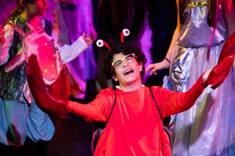 Child in a red crab costume performing on stage, smiling joyfully with arms raised, during a colorful play.