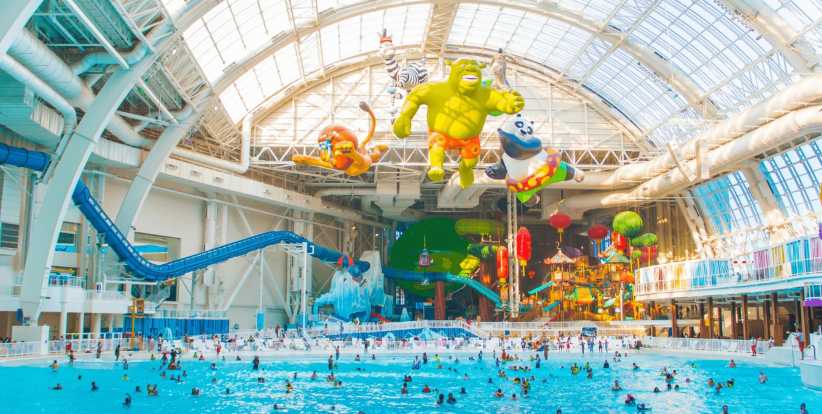 Large inflatable cartoon characters float above a crowded indoor wave pool; water slides and play structures surround the pool within a large, glass-roofed building.