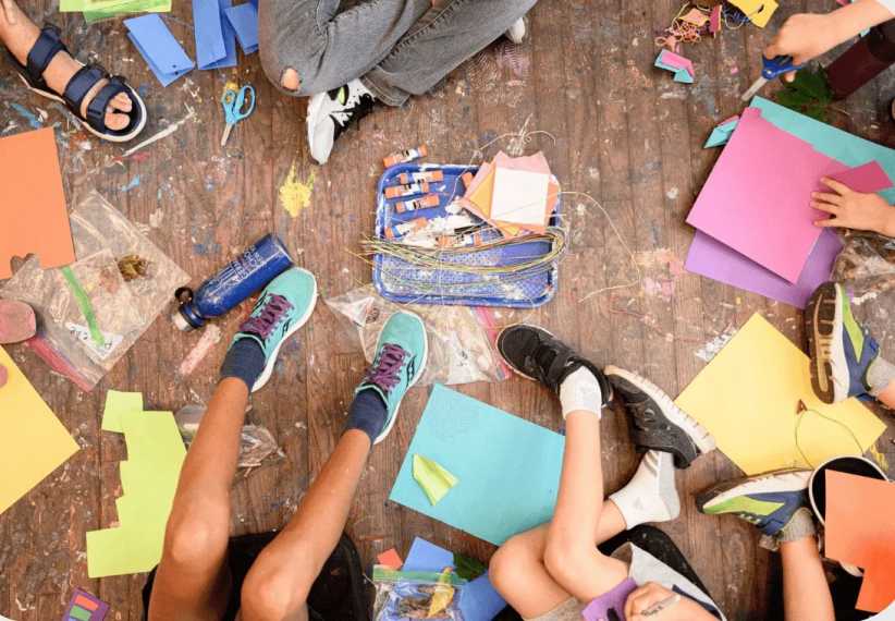 Kids sitting on a wooden floor surrounded by colorful paper, scissors, glue, and craft supplies, engaged in arts and crafts.