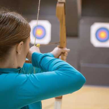A person in a blue shirt aims a wooden bow at two archery targets indoors, focusing intently on their shot.