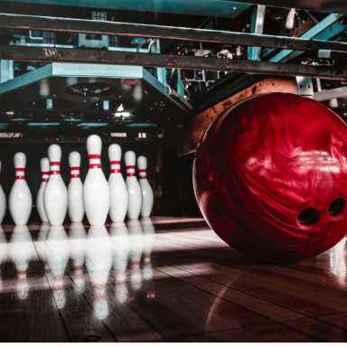 A shiny red bowling ball rolls toward a set of white pins with red stripes on a polished bowling alley lane under mechanical equipment.