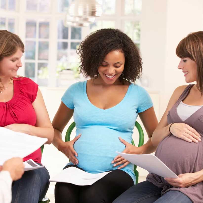 A pregnant woman smiles, holding papers while sitting with two other pregnant women in a bright kitchen.