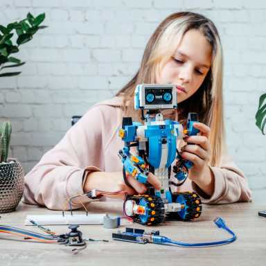 A girl assembles a blue and white Lego robot at a table, surrounded by electronic components and wires. She carefully manipulates small parts.