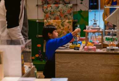 A child wearing a blue shirt interacts with toys or models on a display table, with colorful decorations and adults nearby.