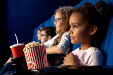 A young girl watches a movie, attentively holding popcorn and a drink, in a dark movie theater alongside other children.
