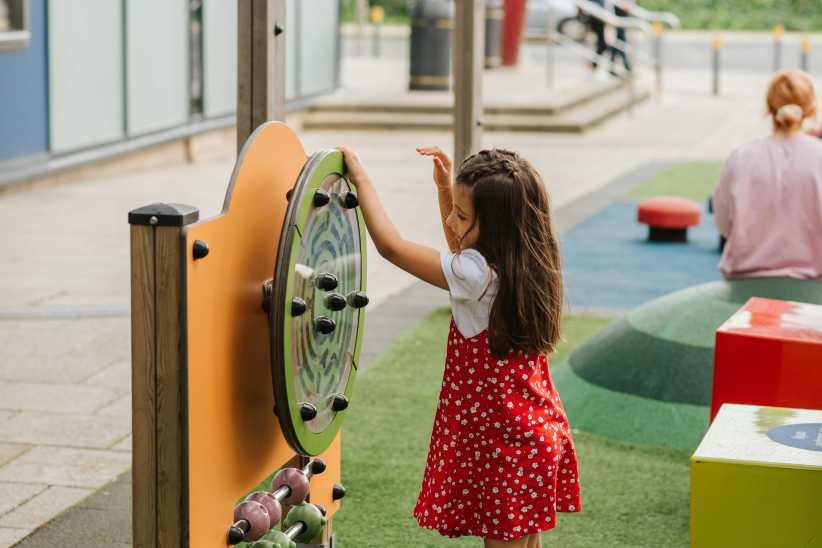 A young girl in a red dress with white flowers plays at an outdoor playground, interacting with a spinning activity station.