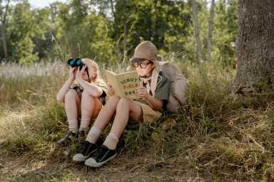 Two children sit on grass in a forest; one reads a "Scout Field Book" while the other looks through binoculars, enjoying nature.