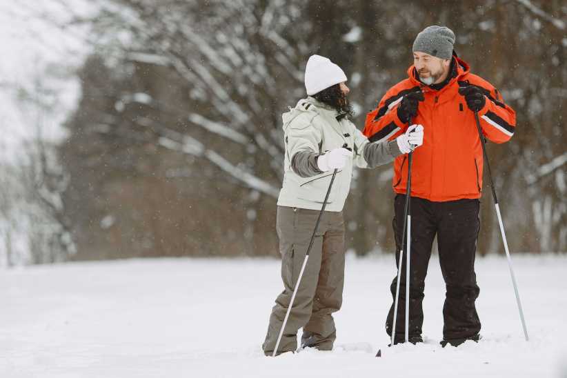 A man in a bright orange jacket and a woman in a white winter hat cross-country skiing together on a snowy trail surrounded by trees.