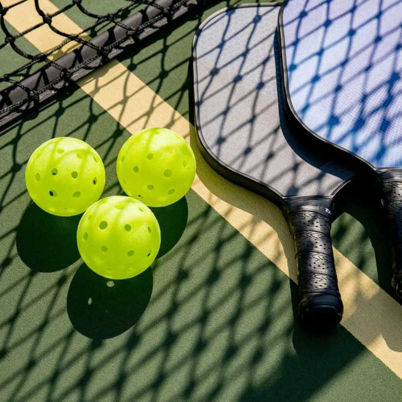Three lime-green pickleballs rest near two pickleball paddles on a court. The paddles and balls cast shadows on the green surface. 