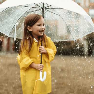 A young girl, wearing a yellow raincoat, joyfully holds a clear umbrella outdoors during a rainstorm.