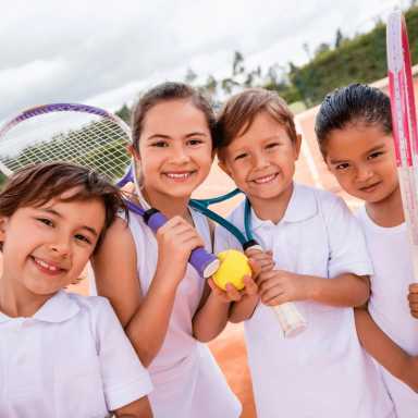 Four smiling children in white shirts hold tennis racquets and a ball, standing together on an outdoor tennis court.