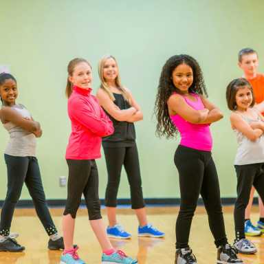 A group of six children—boys and girls—stand confidently in a brightly lit dance studio, each crossing their arms and smiling toward the camera.