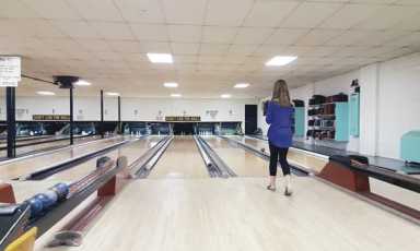 It reveals a retro-style duckpin bowling alley: multiple narrow bowling lanes stretch ahead under a drop ceiling, with a person—seen from behind—rolling a ball, and bowling balls and racks visible to the right.