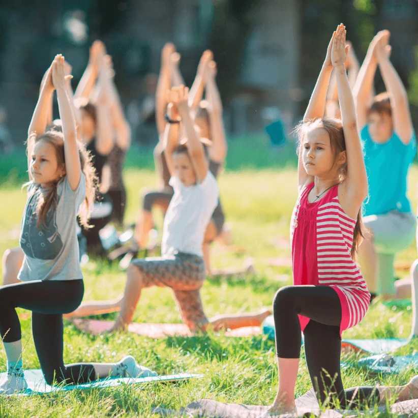 A group of children practice yoga outdoors on a sunny day, kneeling on mats with arms raised, focused and engaged in the activity.