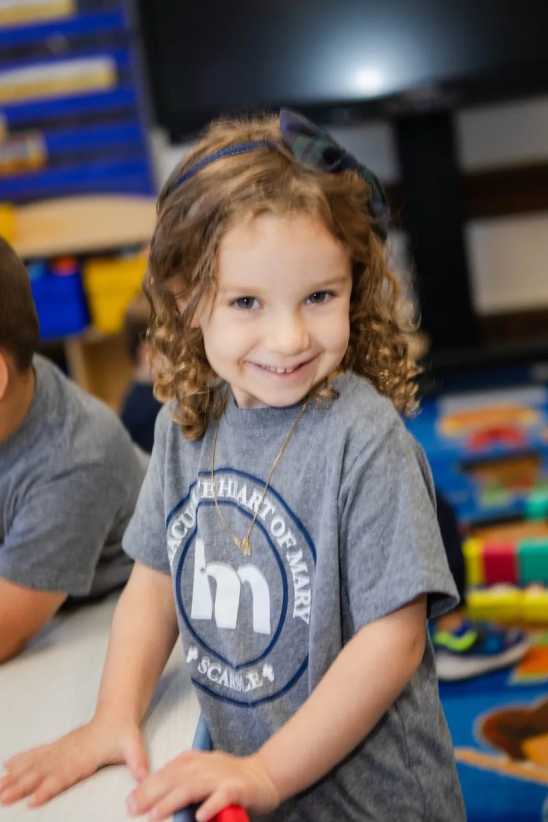 A young child with curly hair in a gray t-shirt stands at a table in a colorful classroom, looking at the camera and smiling.