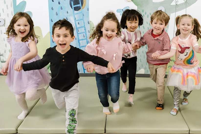 A group of six young children, smiling and holding hands, run toward the camera in a brightly decorated indoor play space.