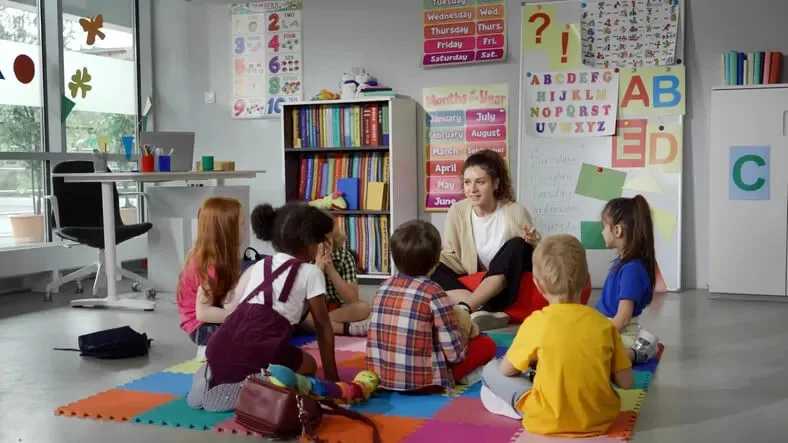 A group of young children sits in a circle on colorful foam mats, listening to a teacher in a bright, decorated classroom.